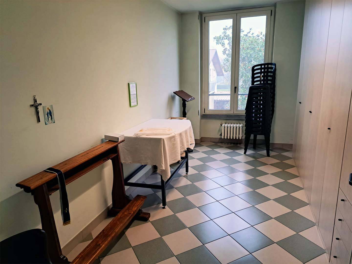 Stanza con finestra adibita a sacrestia in cui sono presenti tavolo, sedie, inginocchiatoio, leggio ed armadio. Appesi al muro crocifisso, immagine della Madonna e quadro
